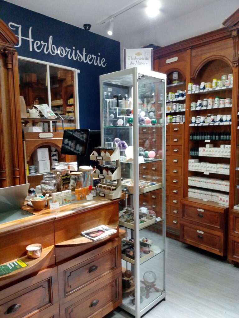 Intérieur de l'Herboristerie du Marais à Paris montrant un comptoir en bois, des étagères remplies de produits naturels, et une vitrine de cristaux.