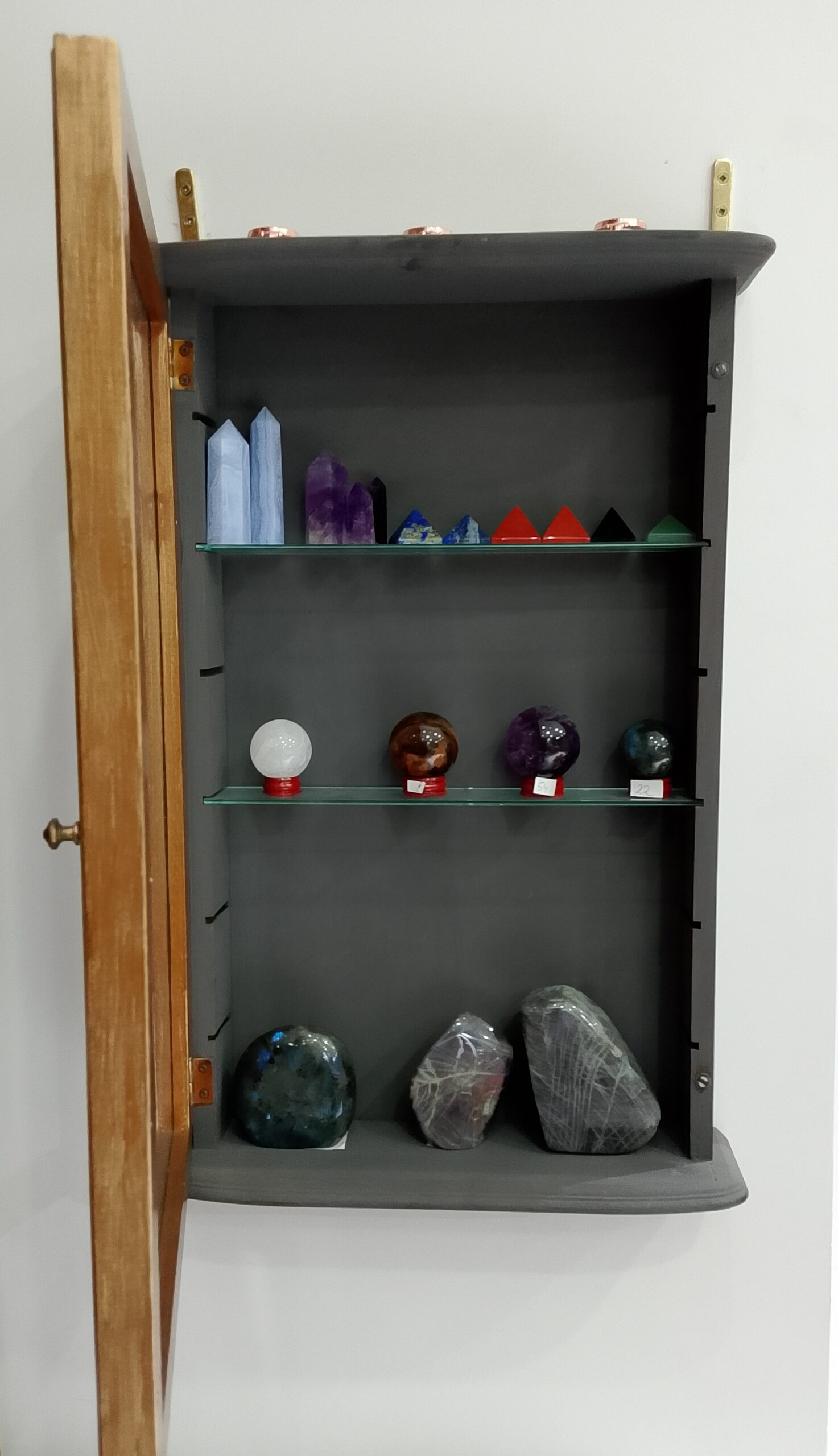 Vitrine murale en bois ouverte montrant des cristaux taillés (pyramides, sphères, amas d'améthyste) sur des étagères en verre.