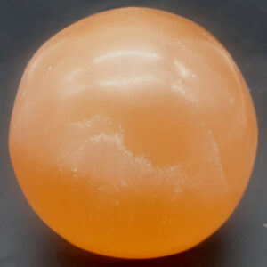 Gros plan sur une pierre sphérique orange translucide posée sur un fond sombre et mat. La surface de la pierre présente une finition lisse et légèrement polie avec des nuances nuageuses et des motifs internes diffus évoquant une texture cristalline naturelle. L'éclairage met en relief l'aspect semi-brillant et les variations de teintes orangées, allant du saumon clair à l'ambre doux.